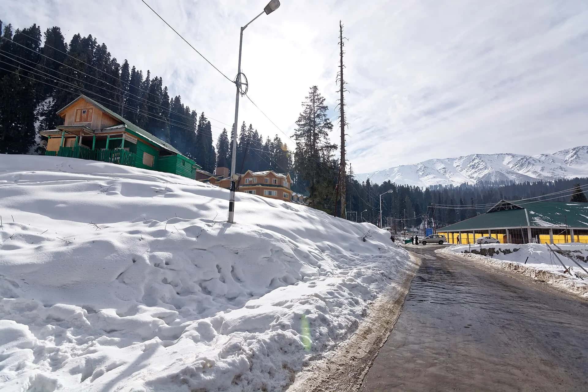 Cleared road through deep snow in Gulmarg, with wooden houses, pine trees and snowy peaks behind