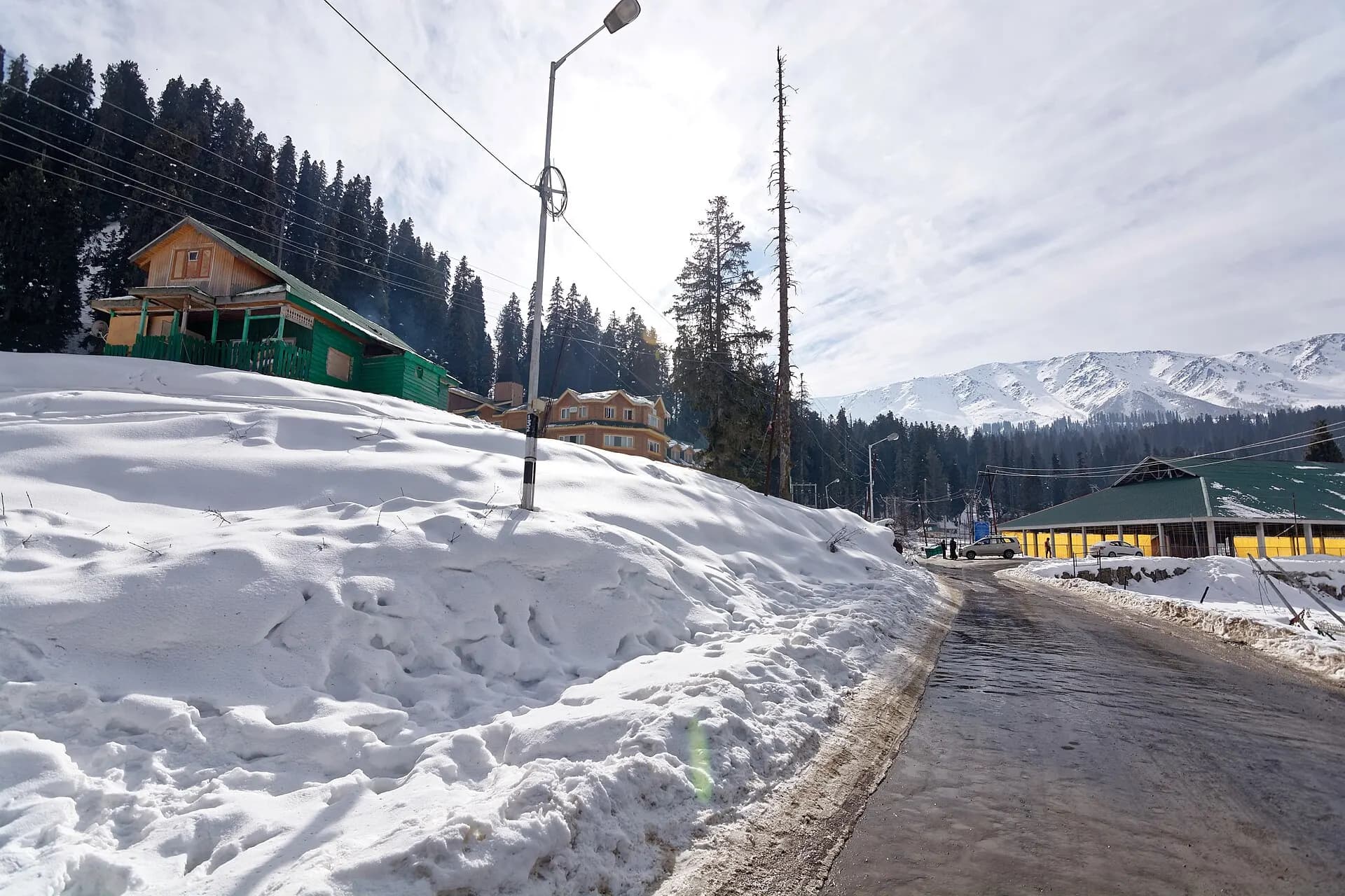Cleared road through deep snow in Gulmarg, with wooden houses, pine trees and snowy peaks behind